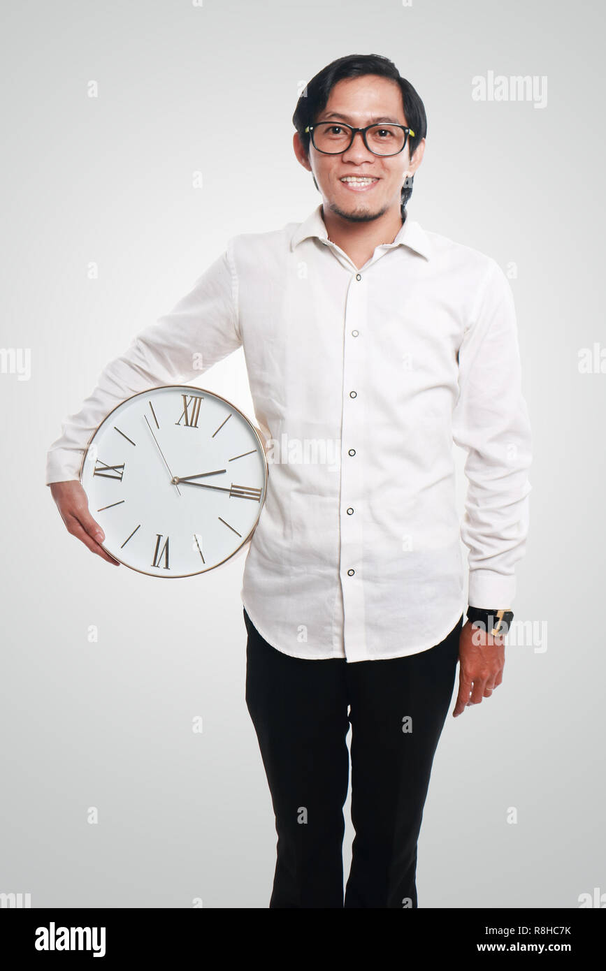 Photo image portrait of a funny young Asian businessman smiling and standing relax while holding a clock on his side, close up portrait, time concept Stock Photo
