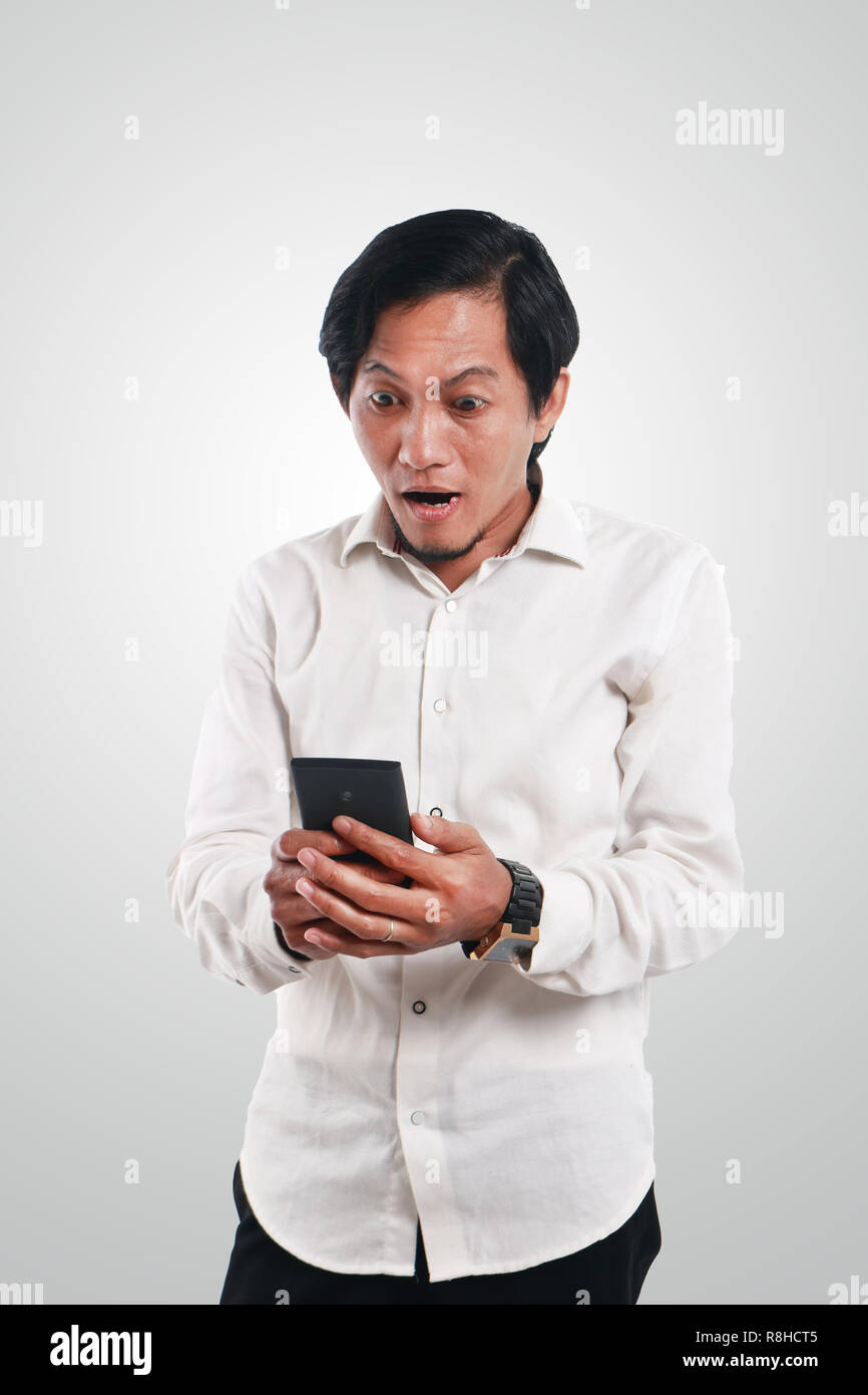 Photo image portrait of a funny young Asian man shocked while looking his smart phone. Holding phone with both hands while reading message on it Stock Photo