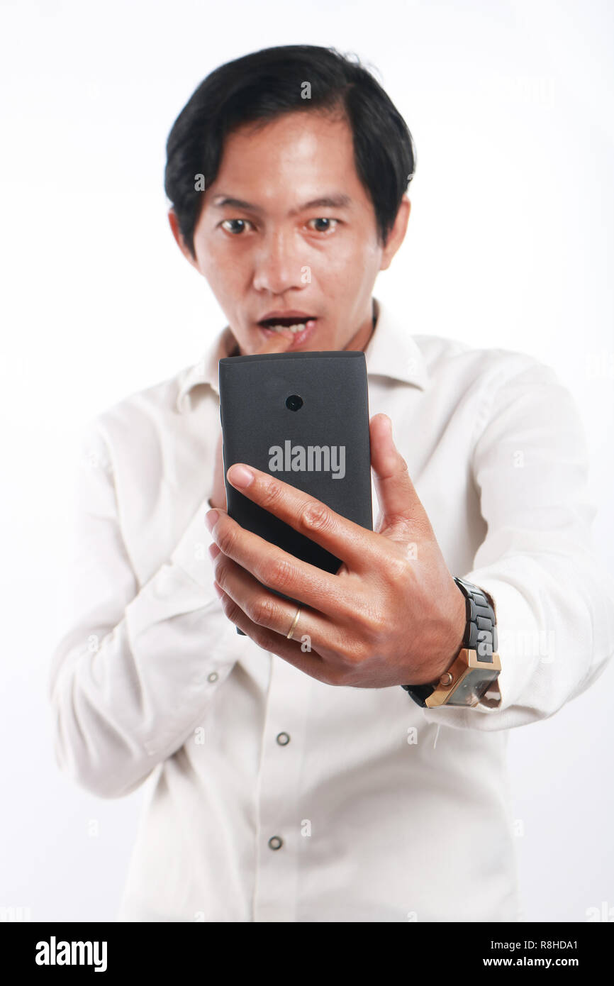 Photo image portrait of a funny young Asian man shocked while looking his smart phone. Holding phone with one hand while reading message on it, over w Stock Photo