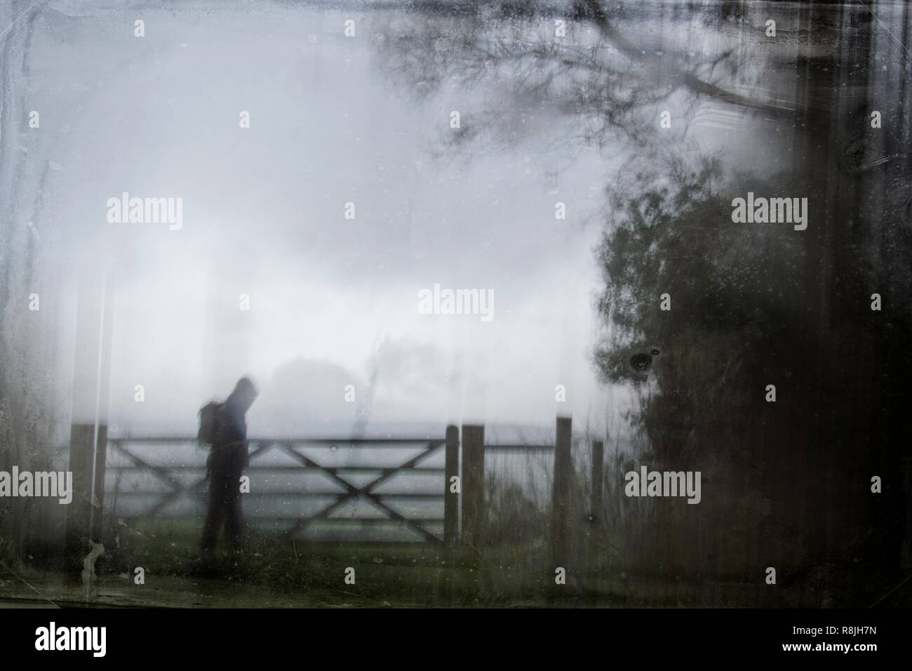 An eerie silhouette of a lone hooded figure with a rucksack by a gate surrounded by trees. With a dark, spooky blurred abstract, grunge effect edit. Stock Photo