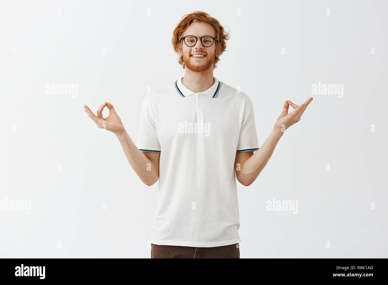 Man is good expample of patience and relaxation. Happy calm good-looking redhead guy with beard in glasses and white casual shirt spreading hands aside in zen gesture meditating and smiling relieved Stock Photo