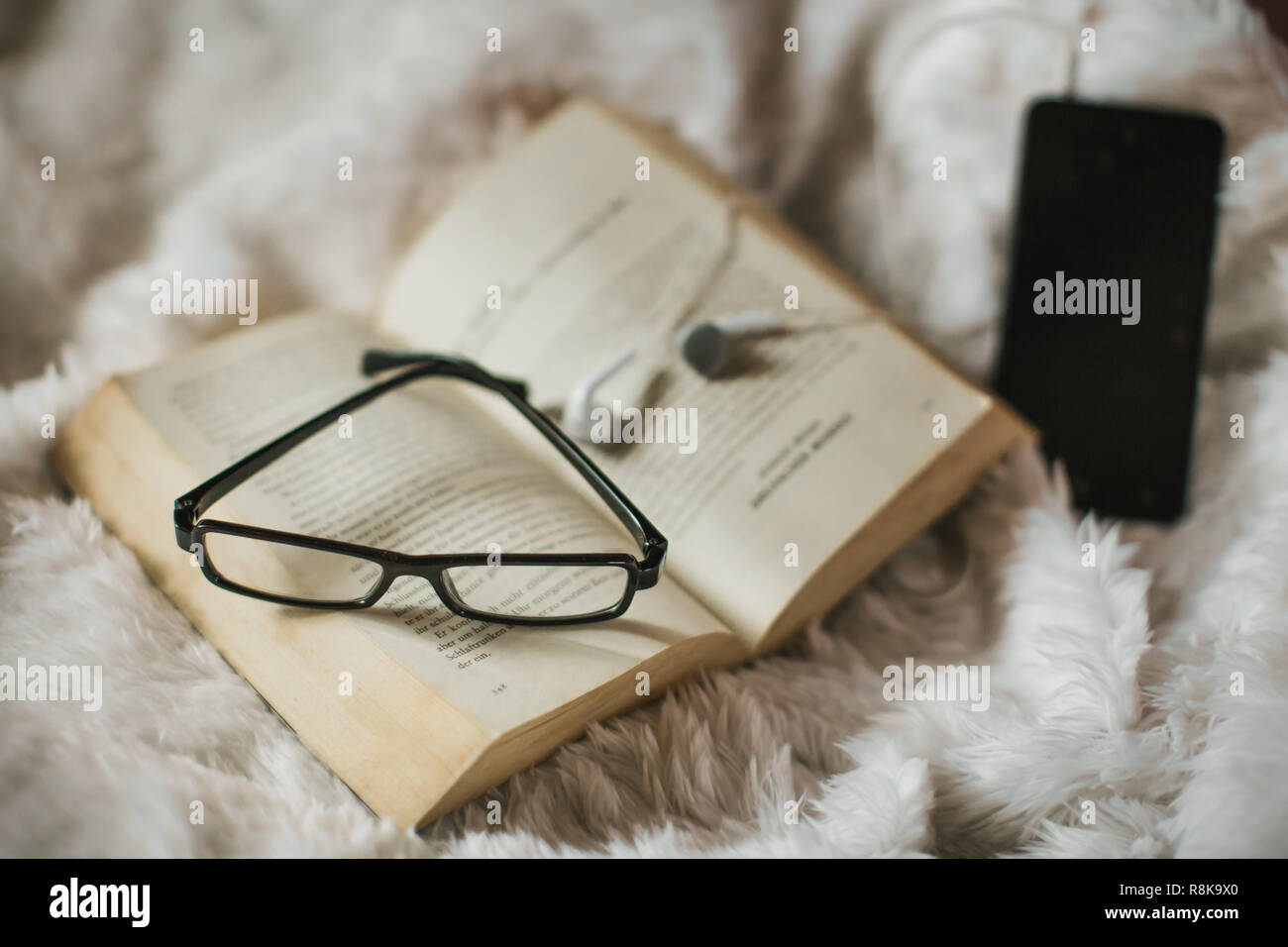an old book on blankie blanket with horn-rimmed eye glasses and a mobile phone with headset, in a peaceful scene Stock Photo