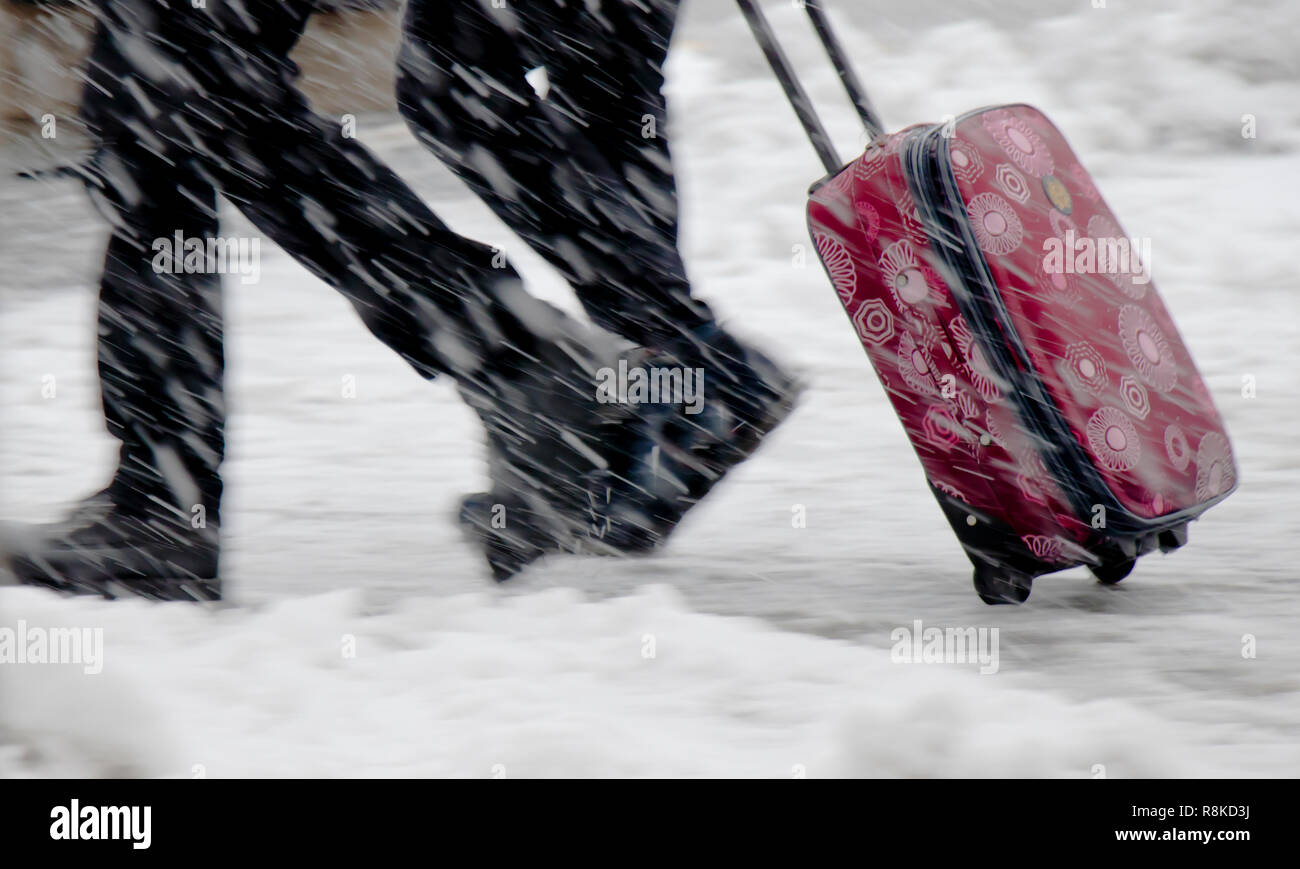 Winter travel; Blurry legs of two young people walking fast in heavy snowfall and rolling red wheeled suitcase Stock Photo