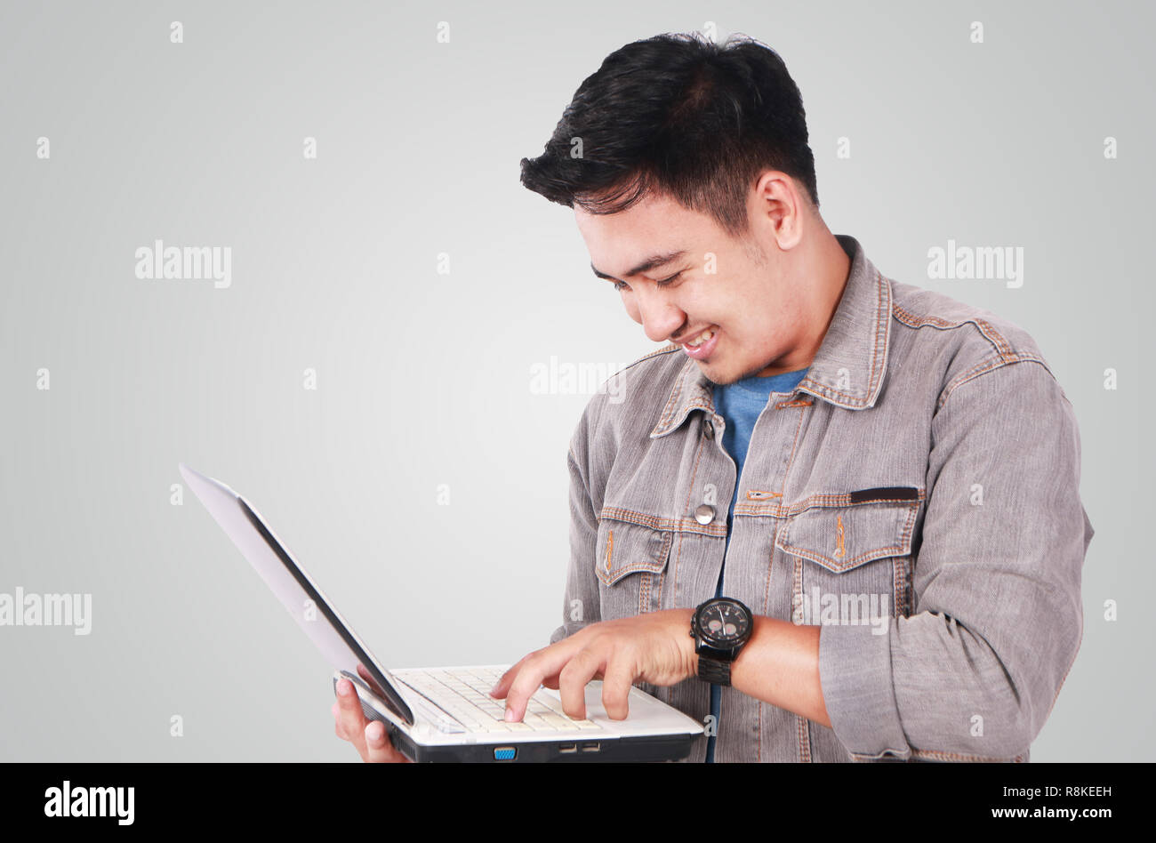 Photo image portrait of a cute young Asian male student standing and smiling while holding laptop and typing on it Stock Photo