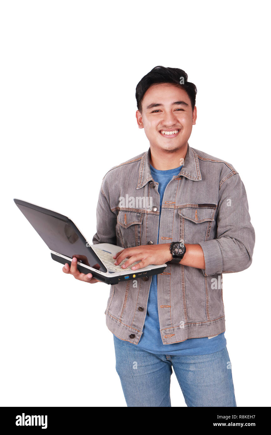 Photo image portrait of a cute young Asian male student standing and smiling while holding laptop and typing on it, isolated on white Stock Photo