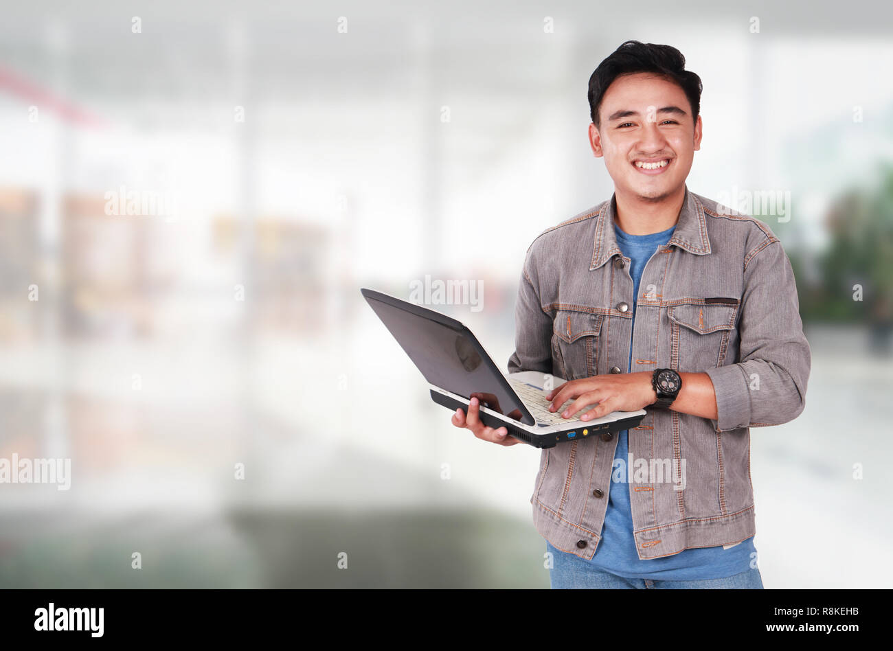 Photo image portrait of a cute young Asian male student standing and smiling while holding laptop and typing on it Stock Photo