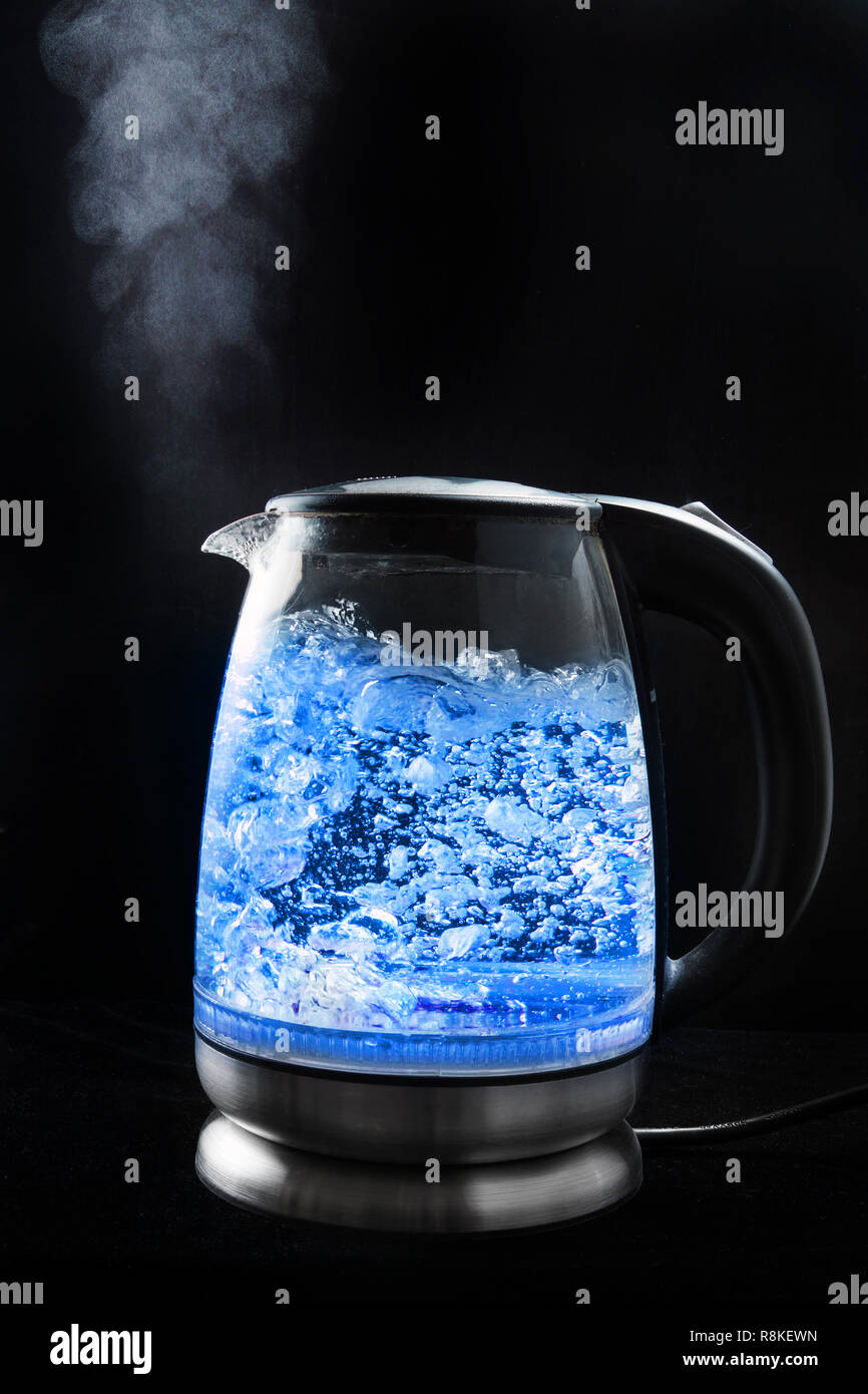Boiling glass kettle with blue light on a black background, steam comes ...