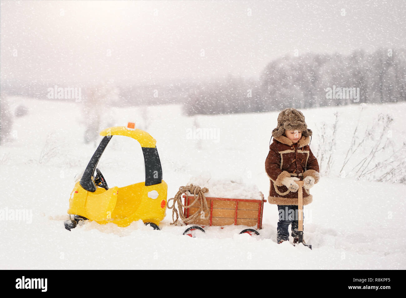 Happy little Boy in winter clothes.  The child is playing outside in the winter. A yellow toy car and a small cart filled with snow, stuck in the snow Stock Photo