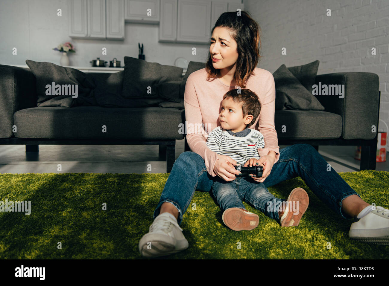 child and his mother sitting on floor and playing video game with joystick at home Stock Photo