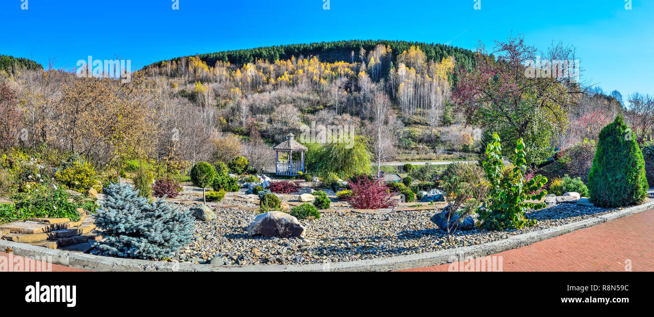 Colorful dwarf coniferous trees, bushes and flowers among stones in autumn garden and wooden arbor in the shade of spreading willows. Beautiful landsc Stock Photo