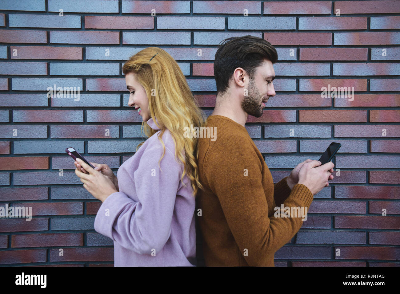 Side portrait of beautiful young couple is using smartphones and smiling, standing back to back against brown brick wall. Stock Photo