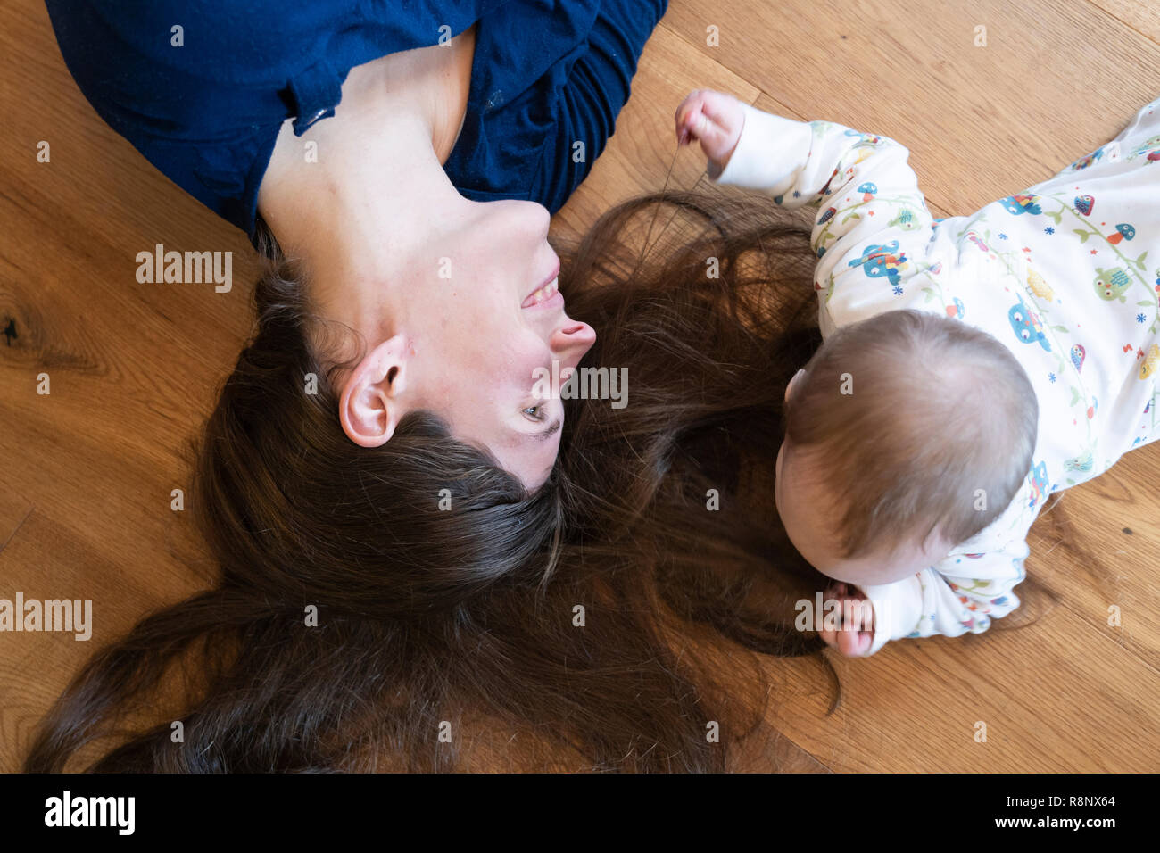 A baby girl lying on the floor and playing with her mother's hair with her mother smiling at her Stock Photo