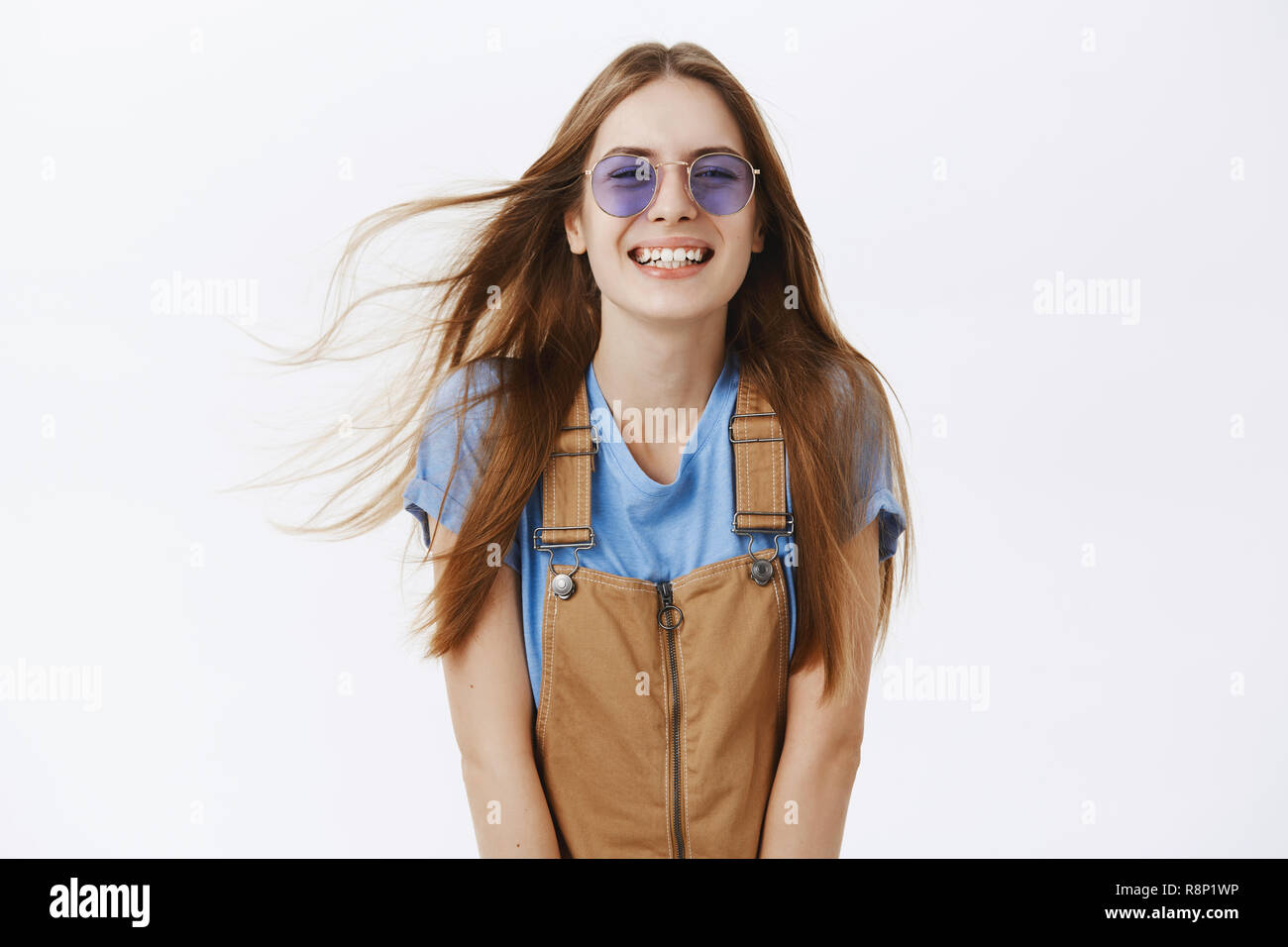 Waist-up shot of charming carefree caucasian girlfriend with long brown hair flicking on air smiling and laughing friendly and playfully posing against gray background in brown overalls and sunglasses Stock Photo