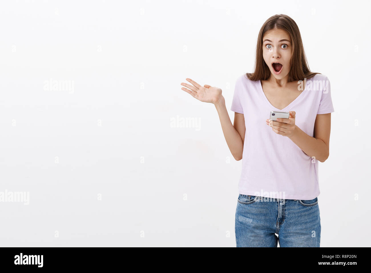 Amazed attractive sociable young female student with long brown hair holding smartphone waving raised palm and dropping jaw from awesome invitation to party received via interned over white wall Stock Photo