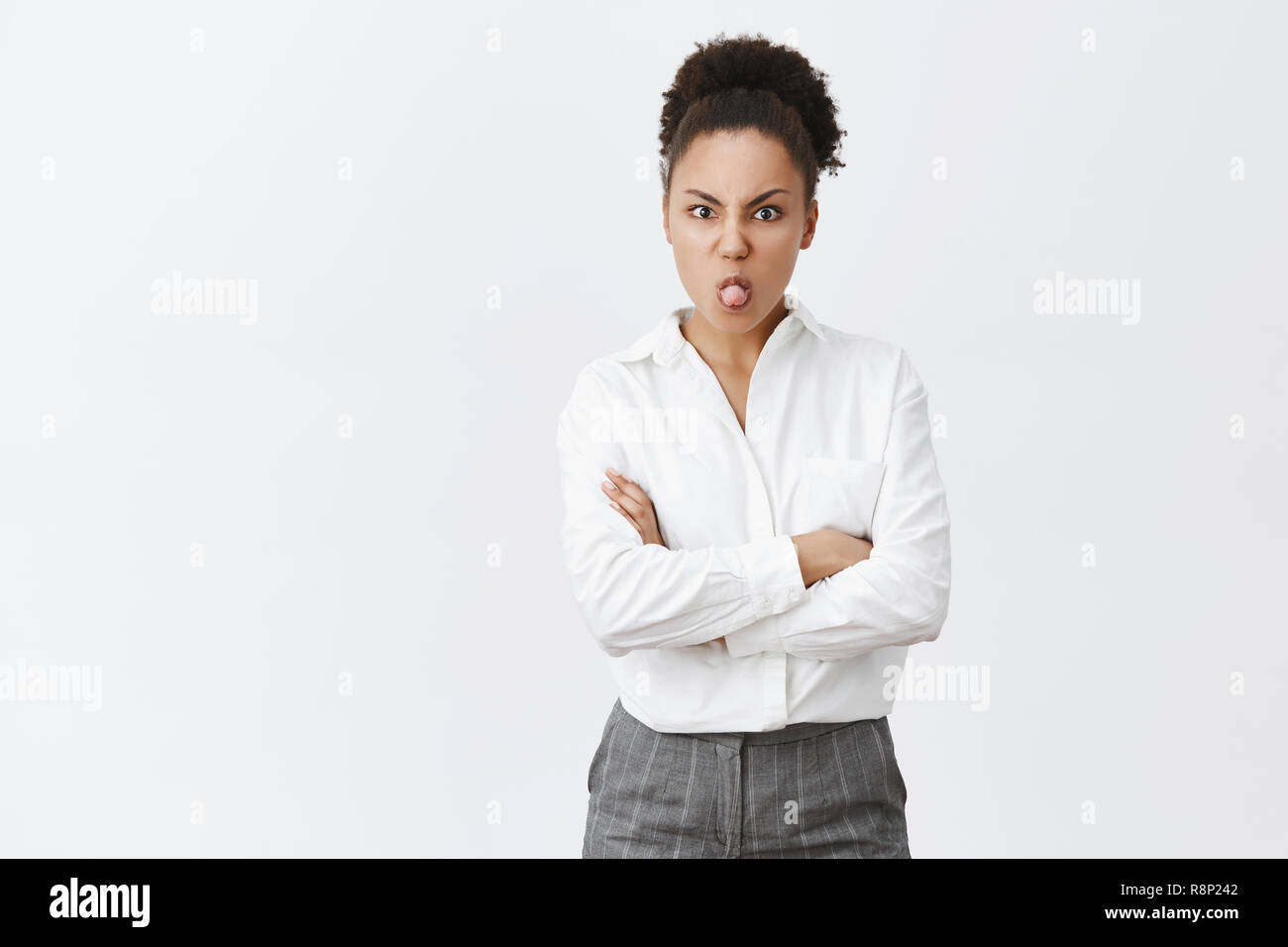 Girl being moody, showing her charachter while having silly argument with girlfriend, holding hands crossed on chest, sticking out tongue and frowning, misbehave and bieng childish over grey wall Stock Photo