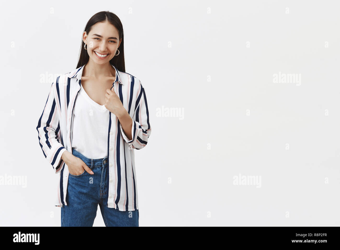 Indoor shot of happy sucessful young female entrepreneur in striped blouse and jeans, holding hand in pocket, smiling joyfully, being pleased after opening own business over gray background Stock Photo