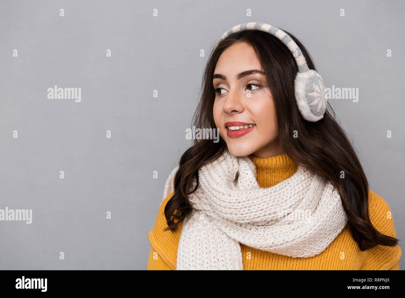 Portrait of optimistic woman wearing ear muffs and scarf smiling and looking aside isolated over gray background Stock Photo