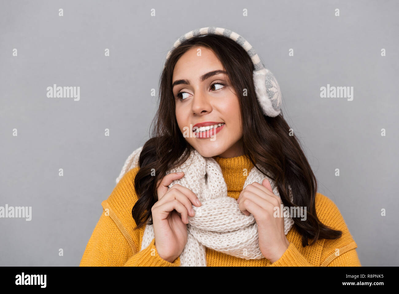 Portrait of beautiful woman wearing ear muffs and scarf smiling and touching scarf isolated over gray background Stock Photo