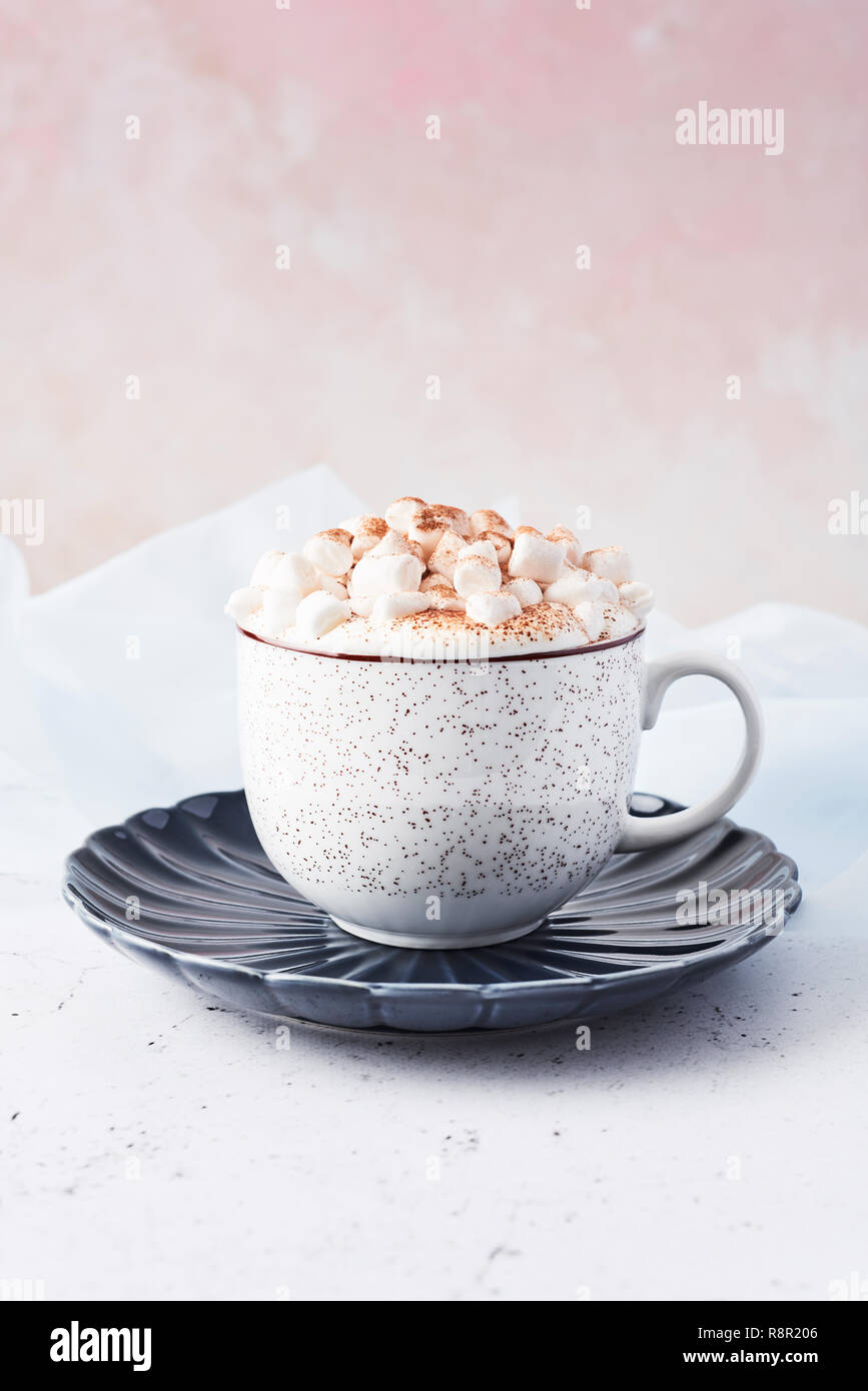 Hot chocolate or cocoa with whipped cream and marshmallow candy sprinkled with cinnamon or cocoa powder on white marble table over blue silk and pink  Stock Photo