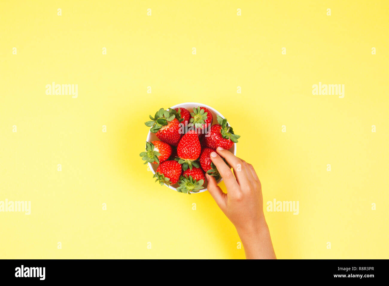Kid hand taking strawberry from the bowl on yellow background. Stock Photo