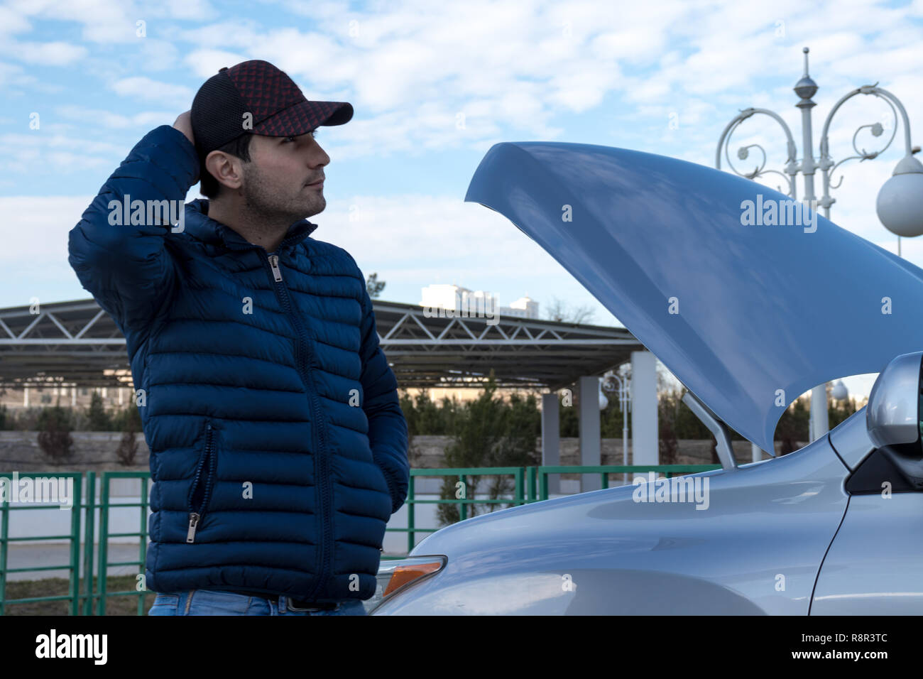 a man thinking how to repair his broken car. Emergency situation. Empty fuel tank. A frustrated and lost young man trying to fix a broken car. Stock Photo