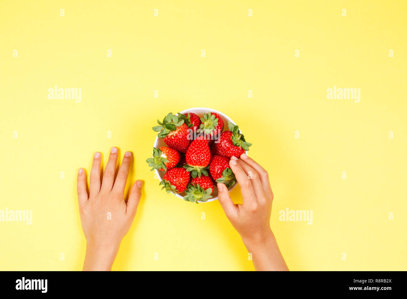 Kid hand taking strawberry from the bowl on yellow background. Stock Photo