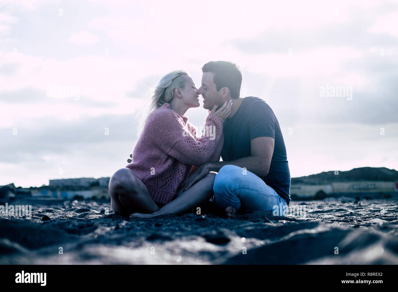Romantic kiss and love with couple in winter enjoying sit down at the beach in nature outdoor scenic place - millennial people lifestyle concept - def Stock Photo