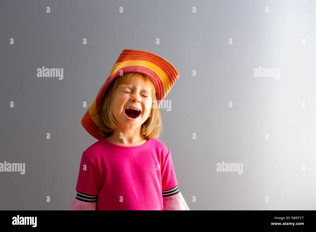 Young girl in pink dress and colorful straw hat screaming or singing with her mouth open wide, standing against grey wall background with copy space.  Stock Photo