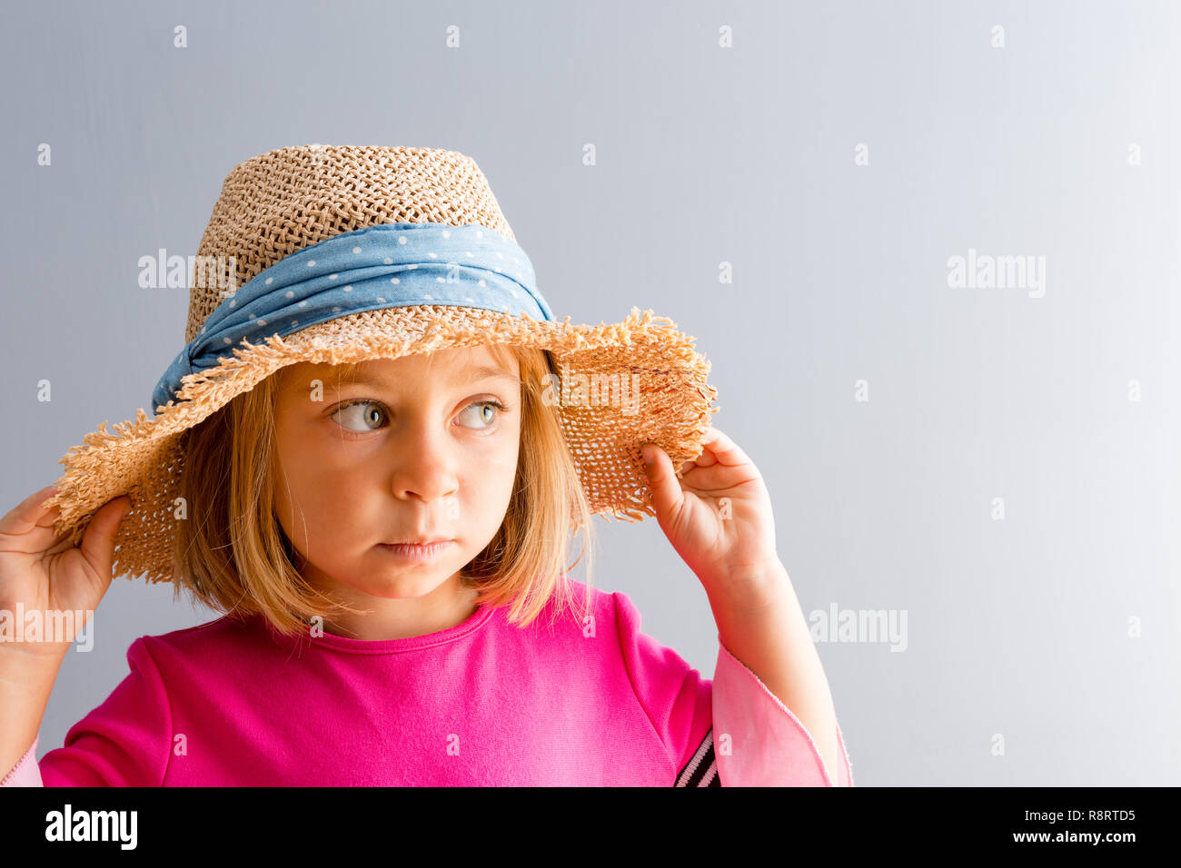 Young pretty girl in pink dress and rural straw hat looking away to the side of copy space on plain grey wall background, while holding her hat with b Stock Photo