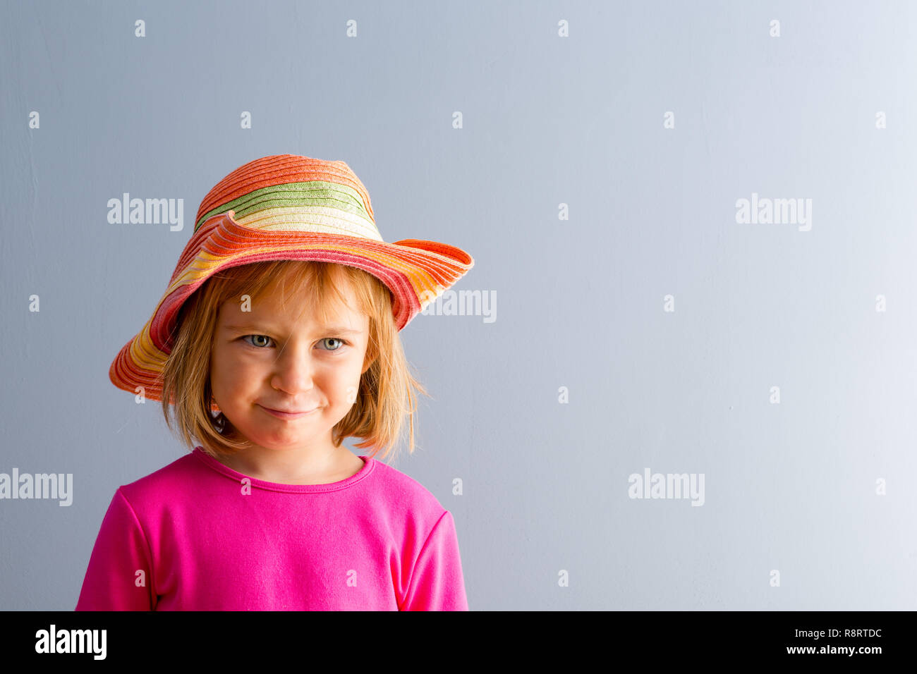 Young blond girl in vivid pink dress and colorful straw hat. Front bust portrait against plain grey background with copy space Stock Photo