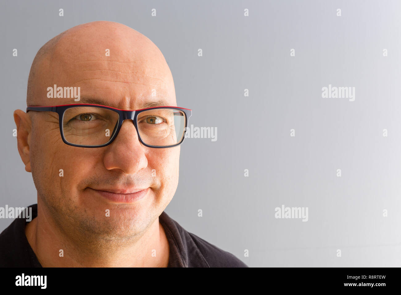 Close-up bust front portrait of smiling bold middle-aged man in black-rimmed glasses and black shirt, looking at camera with a friendly smile. Plain g Stock Photo