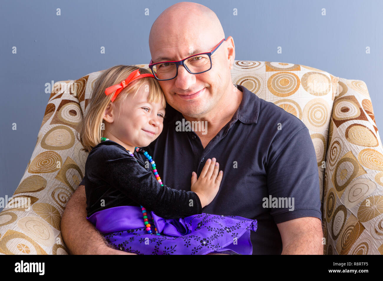 Adult bold man in glasses, father with his happy daughter kid in holiday dressing, sitting in armchair and looking at camera with glad smile. Front ha Stock Photo
