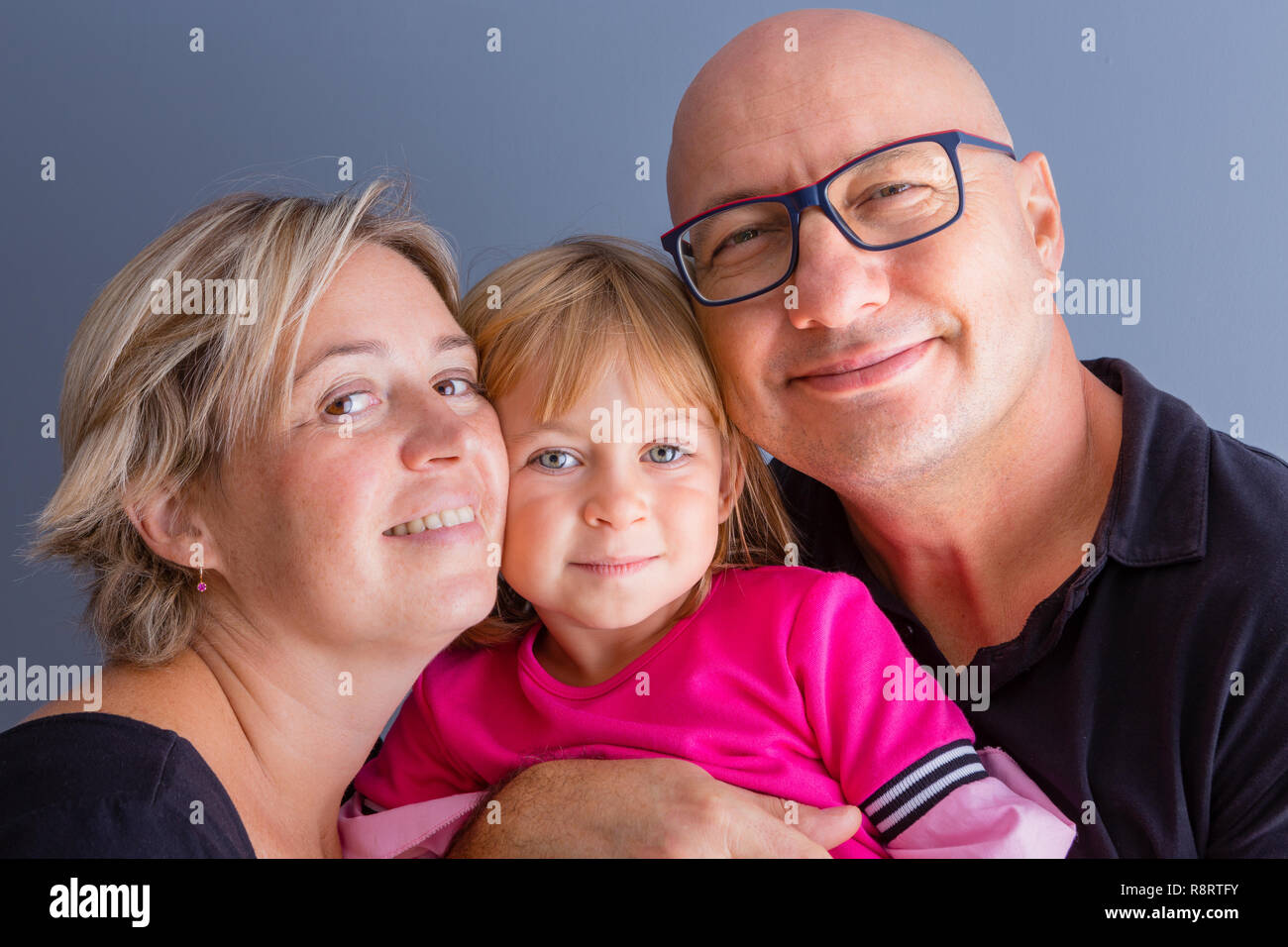 Close-up bust family portrait with parents and young daughter. Bold man in glasses, his blond wife and daughter in pink dress hanging close and smilin Stock Photo