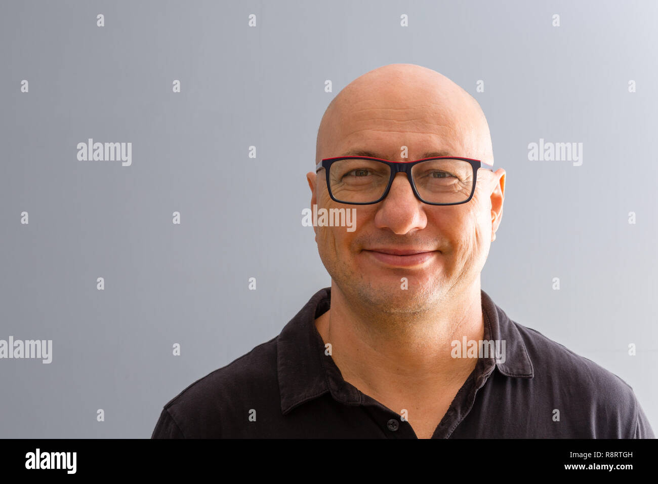 Bust frontal portrait of smiling bold adult man in glasses, wearing black shirt and standing against smooth grey background with copy space Stock Photo