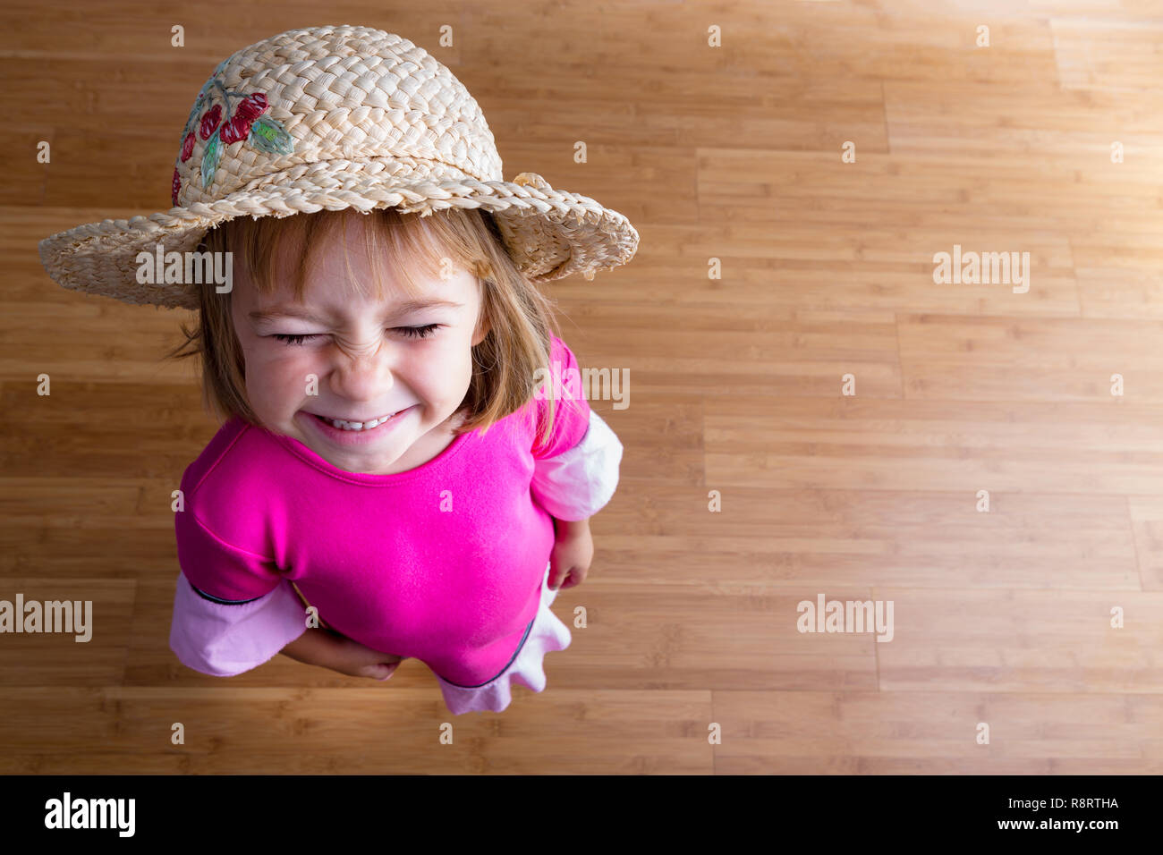 Young girl in straw hat and pink dress, standing on wooden parquet floor and squinting at camera, viewed from above with copy space Stock Photo