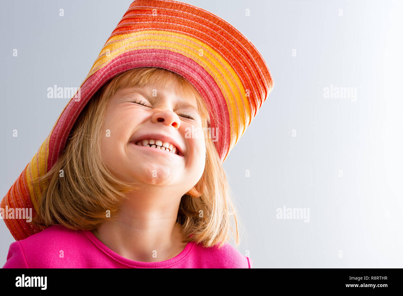 Young girl in colorful hat squinting her face, burst in laughing or crying, bust portrait from low angle, against white background with copy space Stock Photo