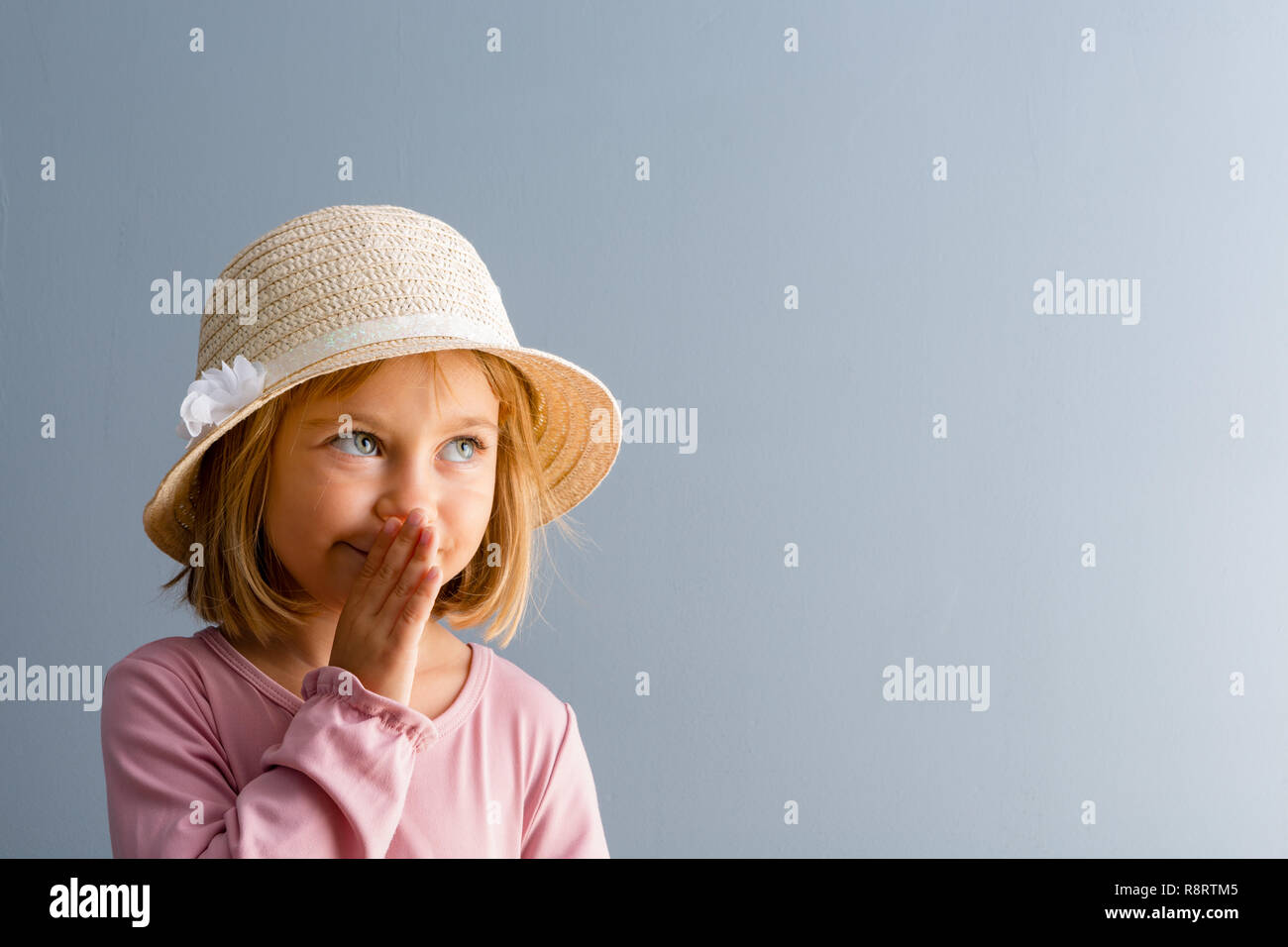 Adorable little four year old girl wearing an elegant straw hat whispering a secret with her hand to her mouth over blue with copy space Stock Photo