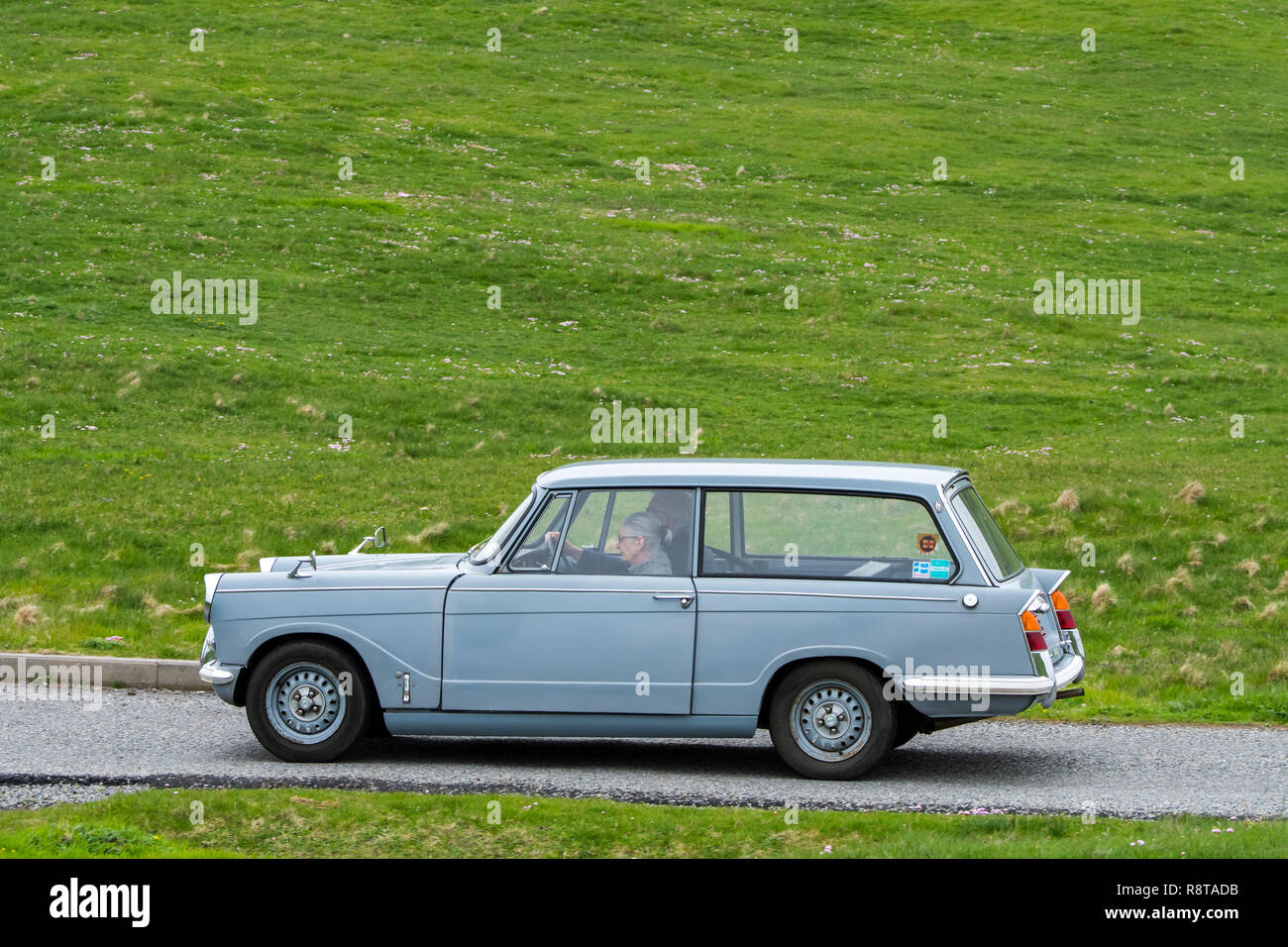 Elderly couple driving 1967 classic car Triumph Herald 1200 Estate along country road through meadow Stock Photo