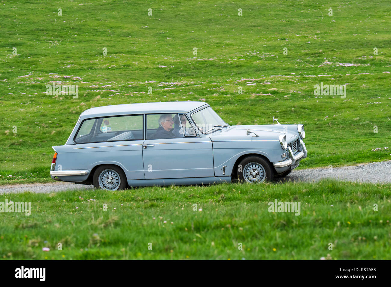 Elderly couple driving 1967 classic car Triumph Herald 1200 Estate along country road through meadow Stock Photo