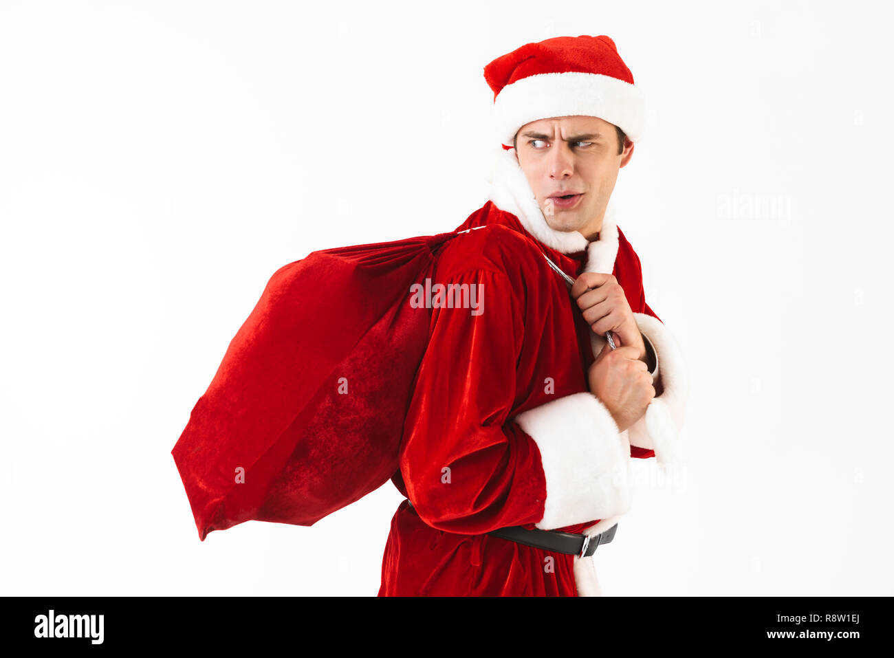 Portrait of displeased man 30s in santa claus costume and red hat walking with gift bag over shoulder isolated on white background in studio Stock Photo