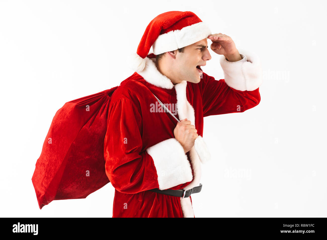 Portrait of handsome man 30s in santa claus costume and red hat walking with gift bag over shoulder isolated on white background in studio Stock Photo