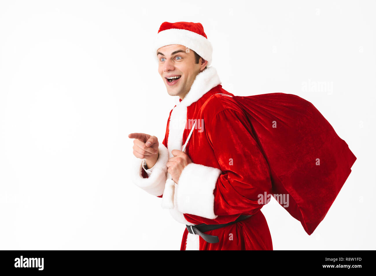 Portrait of joyous man 30s in santa claus costume and red hat walking with gift bag over shoulder isolated on white background in studio Stock Photo