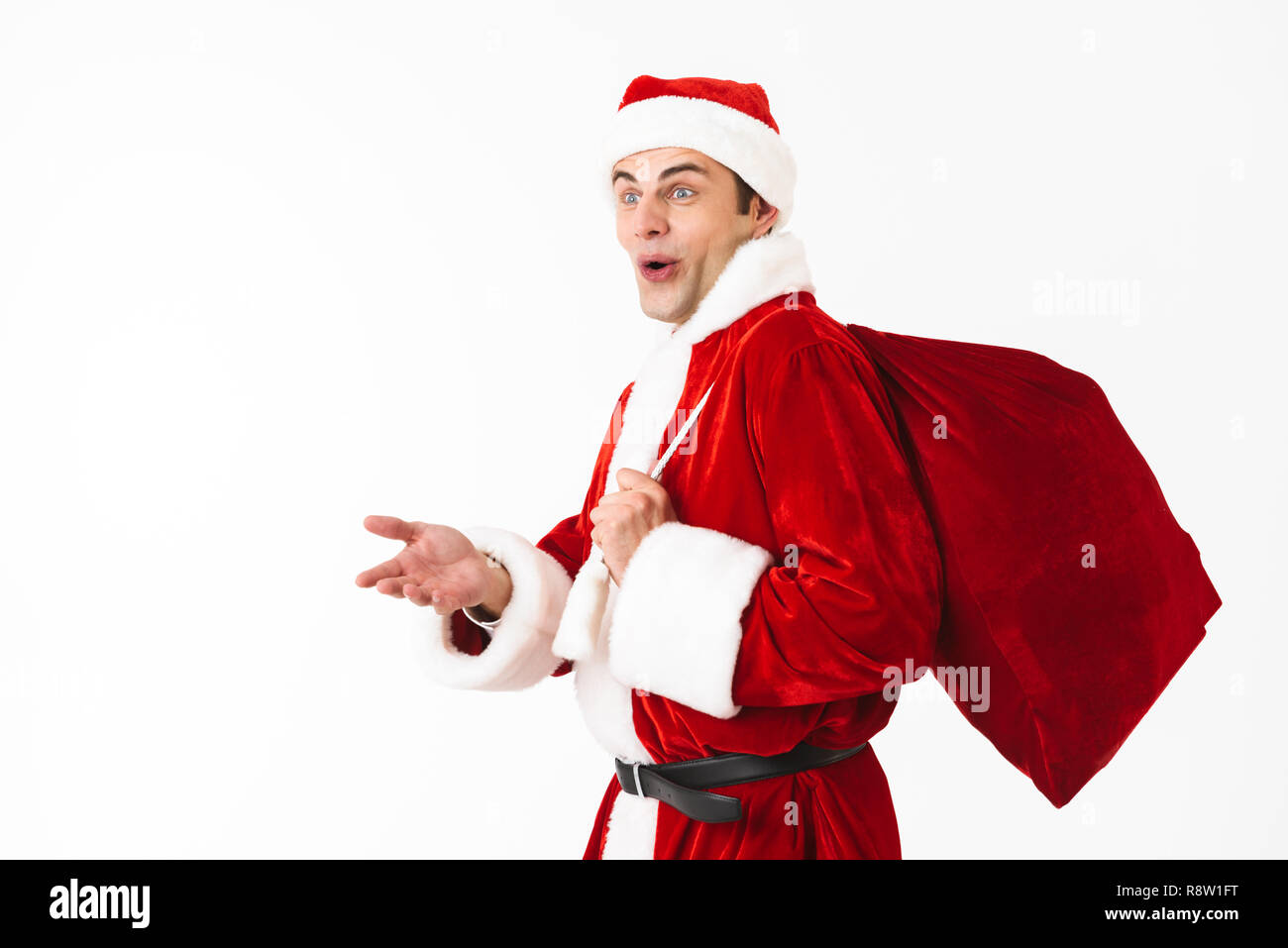Portrait of excited man 30s in santa claus costume and red hat walking with gift bag over shoulder isolated on white background in studio Stock Photo
