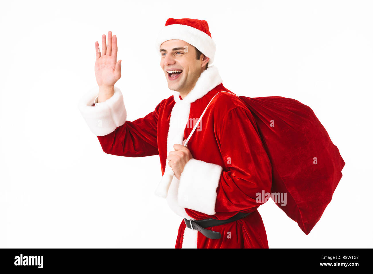 Portrait of caucasian man 30s in santa claus costume and red hat walking with gift bag over shoulder isolated on white background in studio Stock Photo