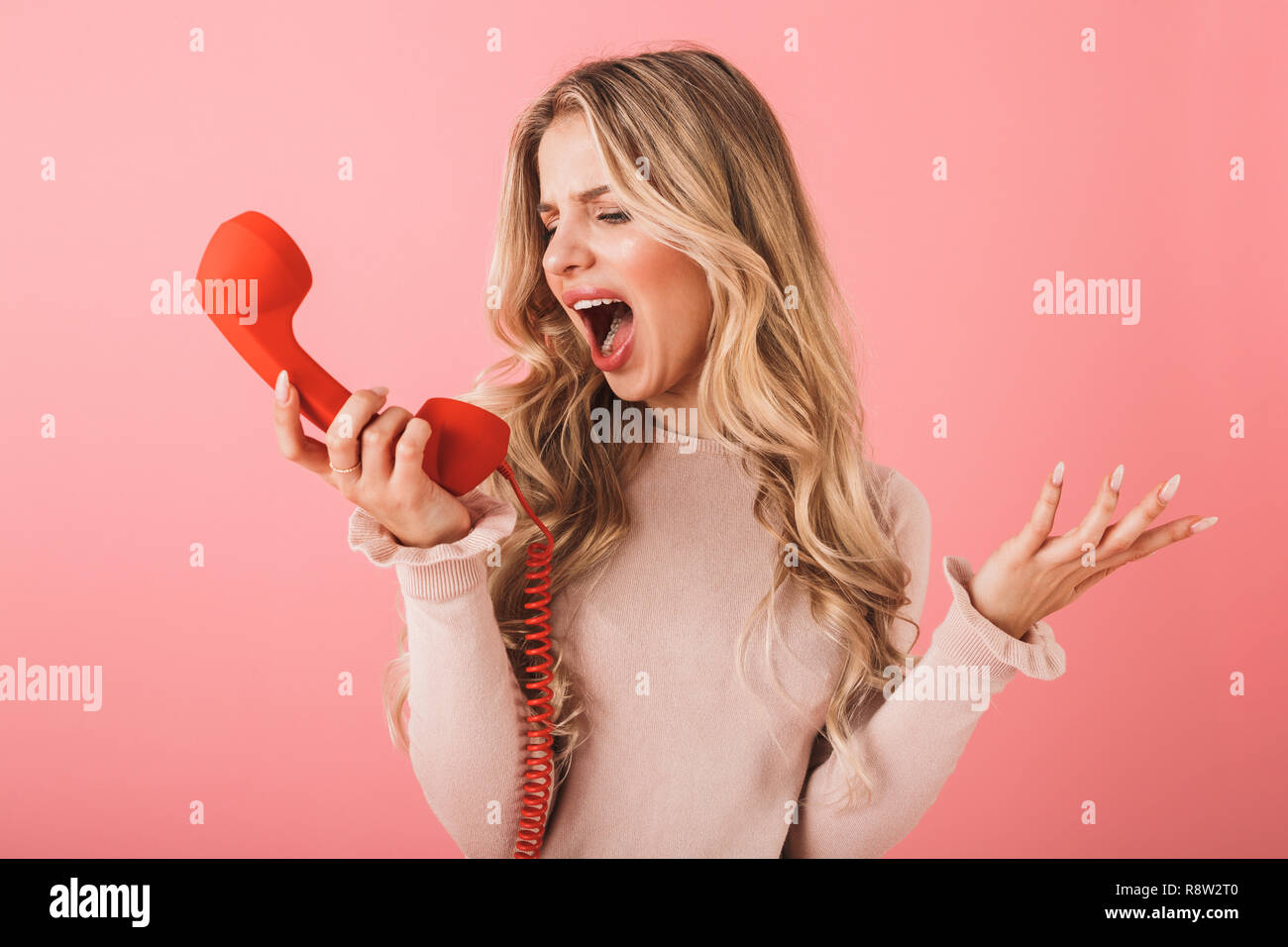 Portrait of an angry blonde haired young woman standing isolated over pink background, talking on a landline phone, yelling Stock Photo
