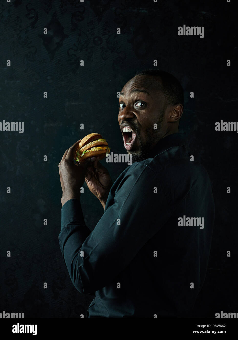 The young african american man eating hamburger and looking away on black studio background Stock Photo