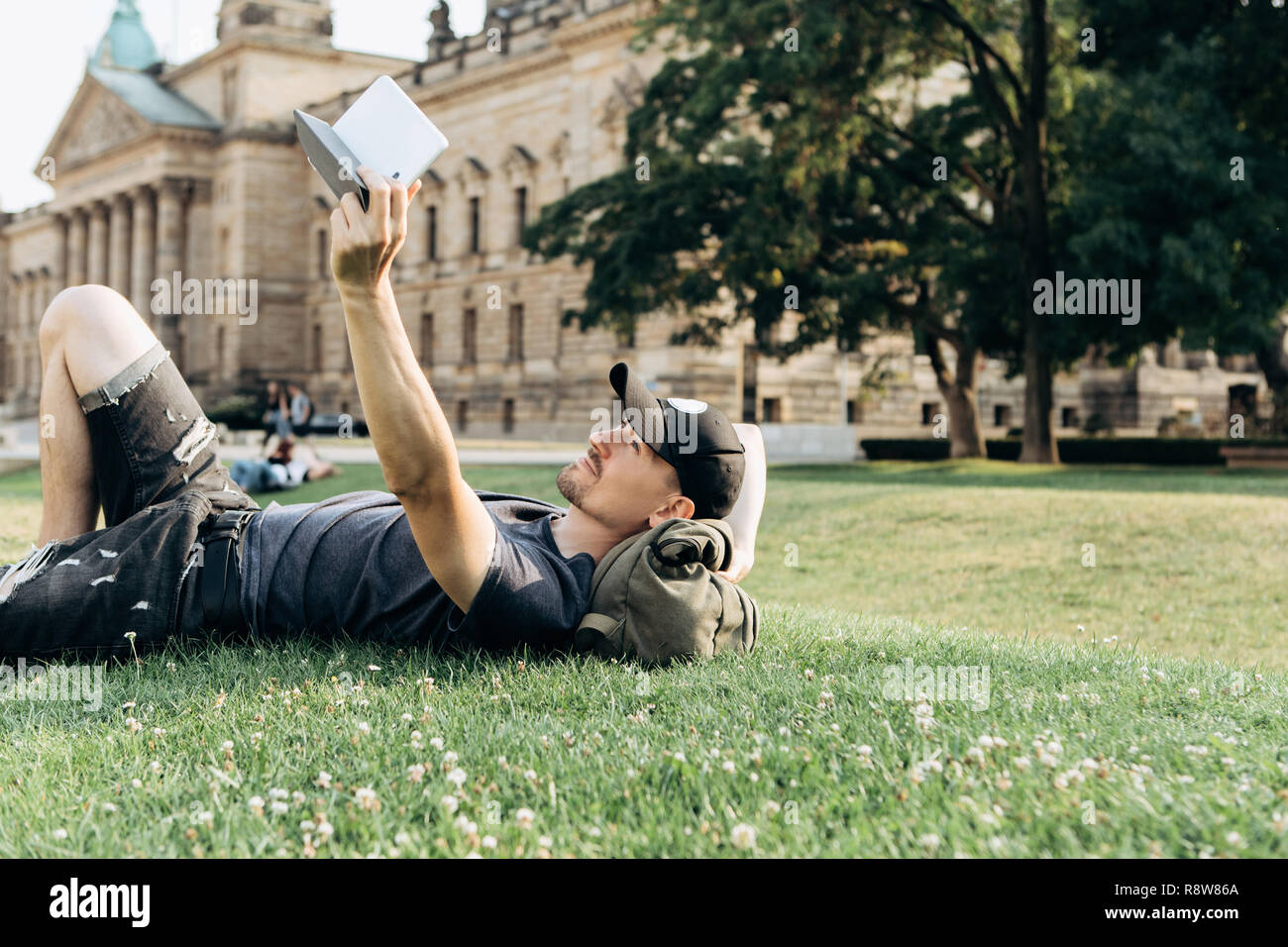 A young guy with a backpack or a student is lying on the grass during a break or is resting and using a tablet. He is watching a video or reading an e-book or something else. Stock Photo