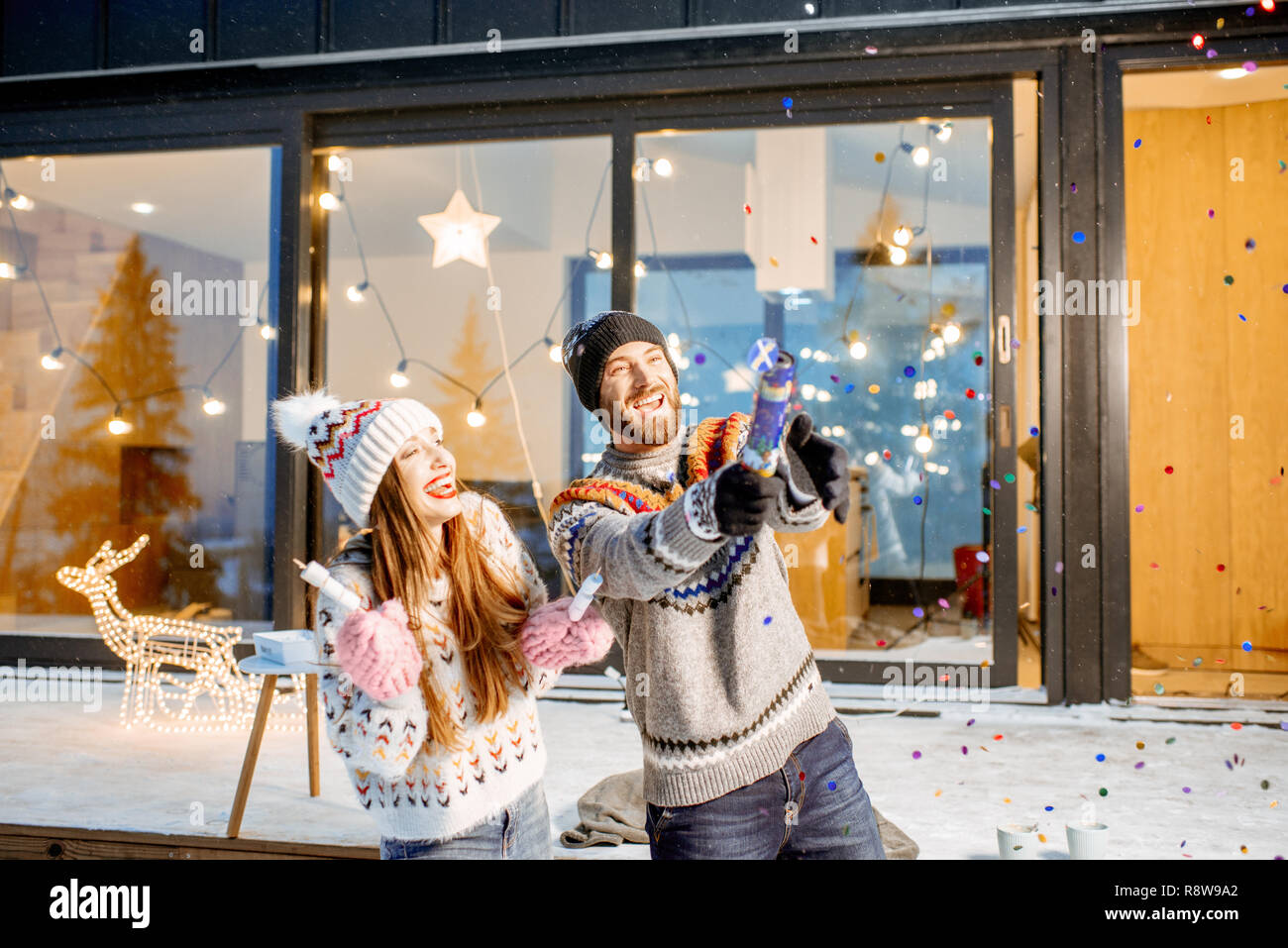 Young happy couple dressed in sweaters celebrating winter holidays in front of a beautiful decorated house in the mountains Stock Photo