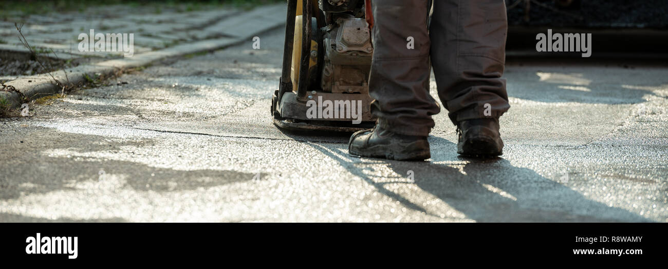 Low angle view of worker using compactor to finish up patching a bump in the road with fresh new asphalt. Wide view image. Stock Photo