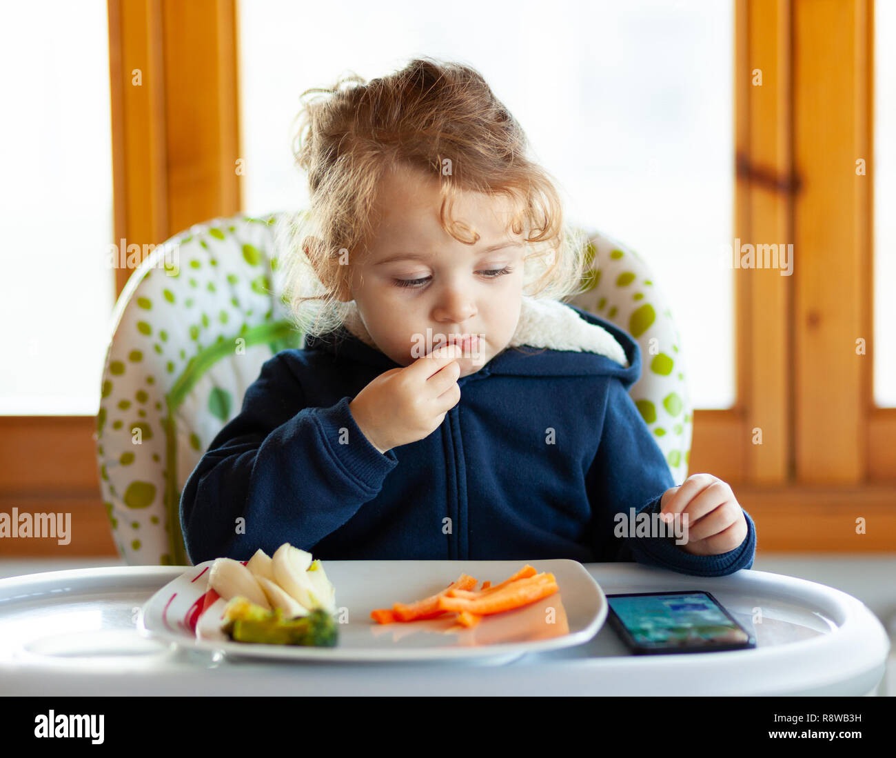 Toddler eats in the high chair while watching movies on the mobile phone. Many children do not want to eat if they do not watch the mobile device or t Stock Photo